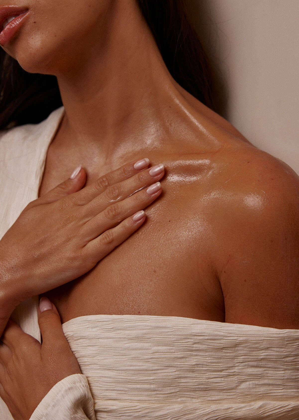 Oiled shoulder and collarbone with hand resting on chest, manicured nails and off-white textured fabric draped across torso.