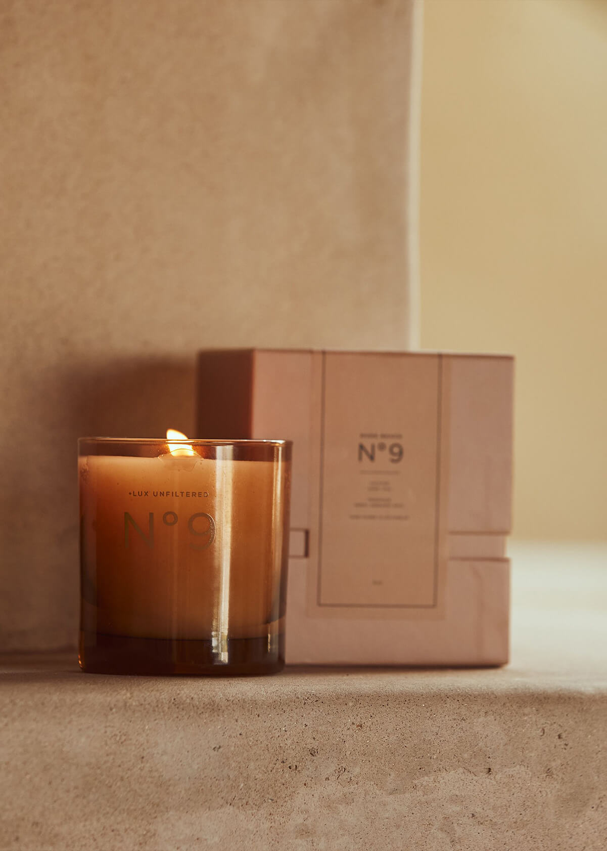 lit amber candle in glass labeled '+LUX UNFILTERED N°9' beside matching boxed N°9 packaging on a stone shelf.