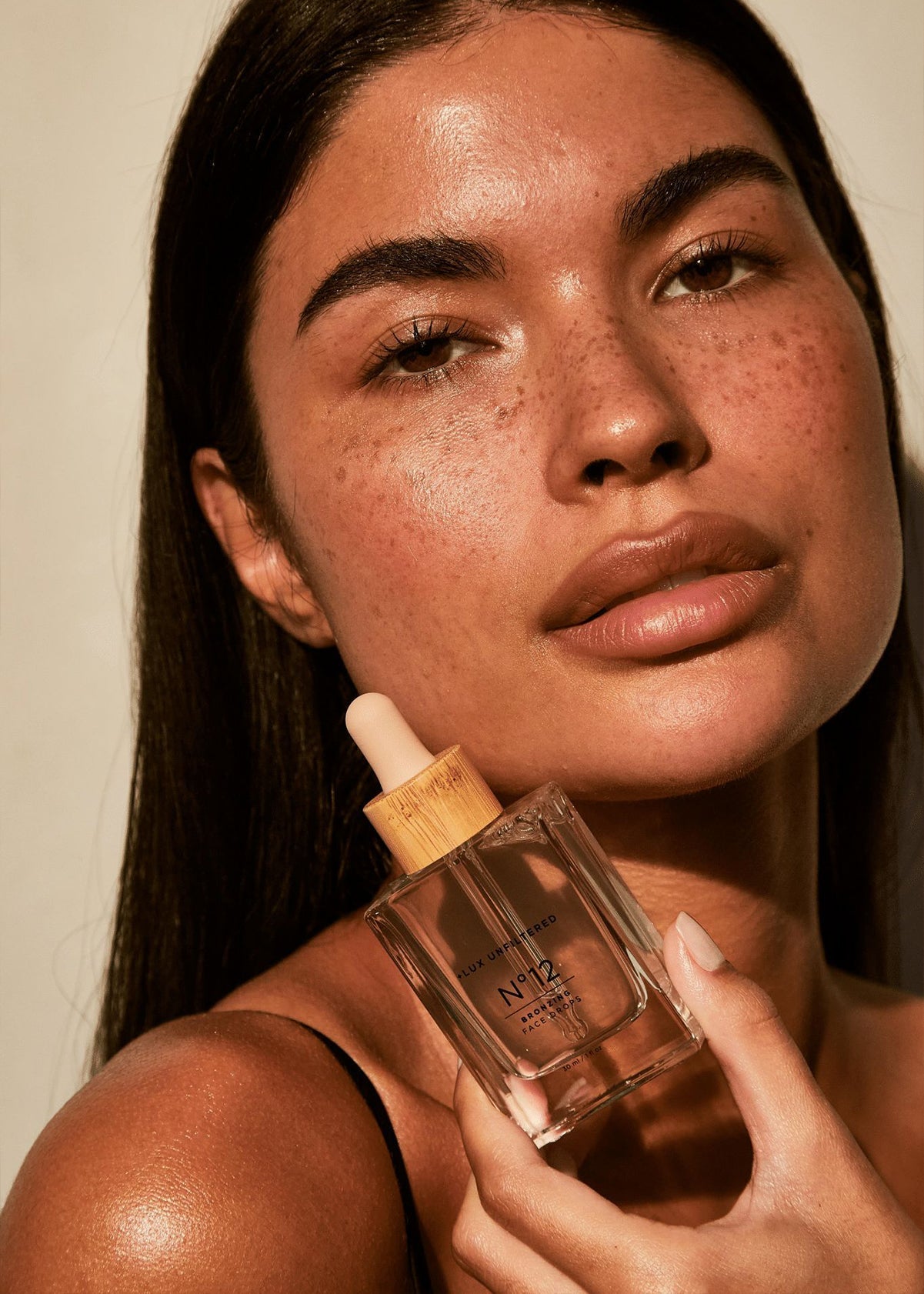 Freckled model holding Lux Unfiltered N°12 bronzing face drops bottle near her cheek.