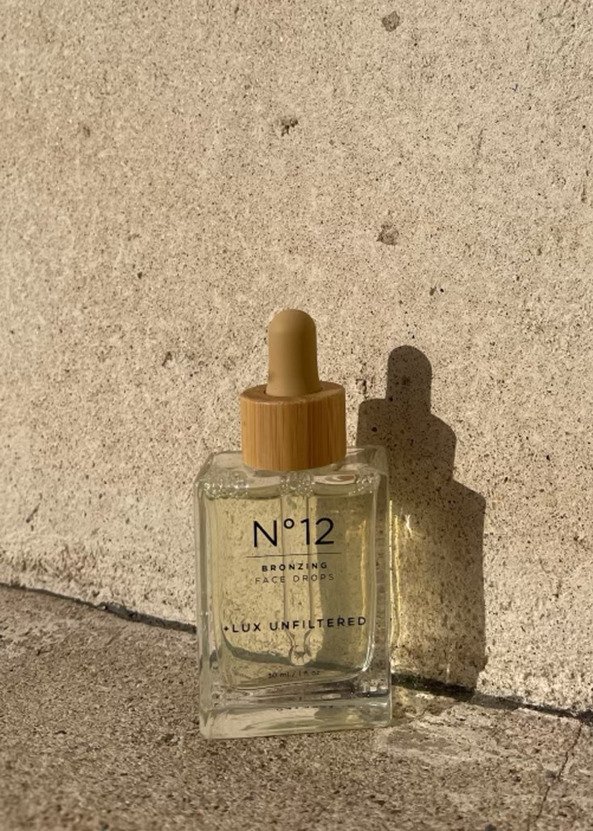 Glass bottle labeled N°12 BRONZING FACE DROPS LUX UNFILTERED, 30 ml / 1 fl oz, wooden dropper casting shadow