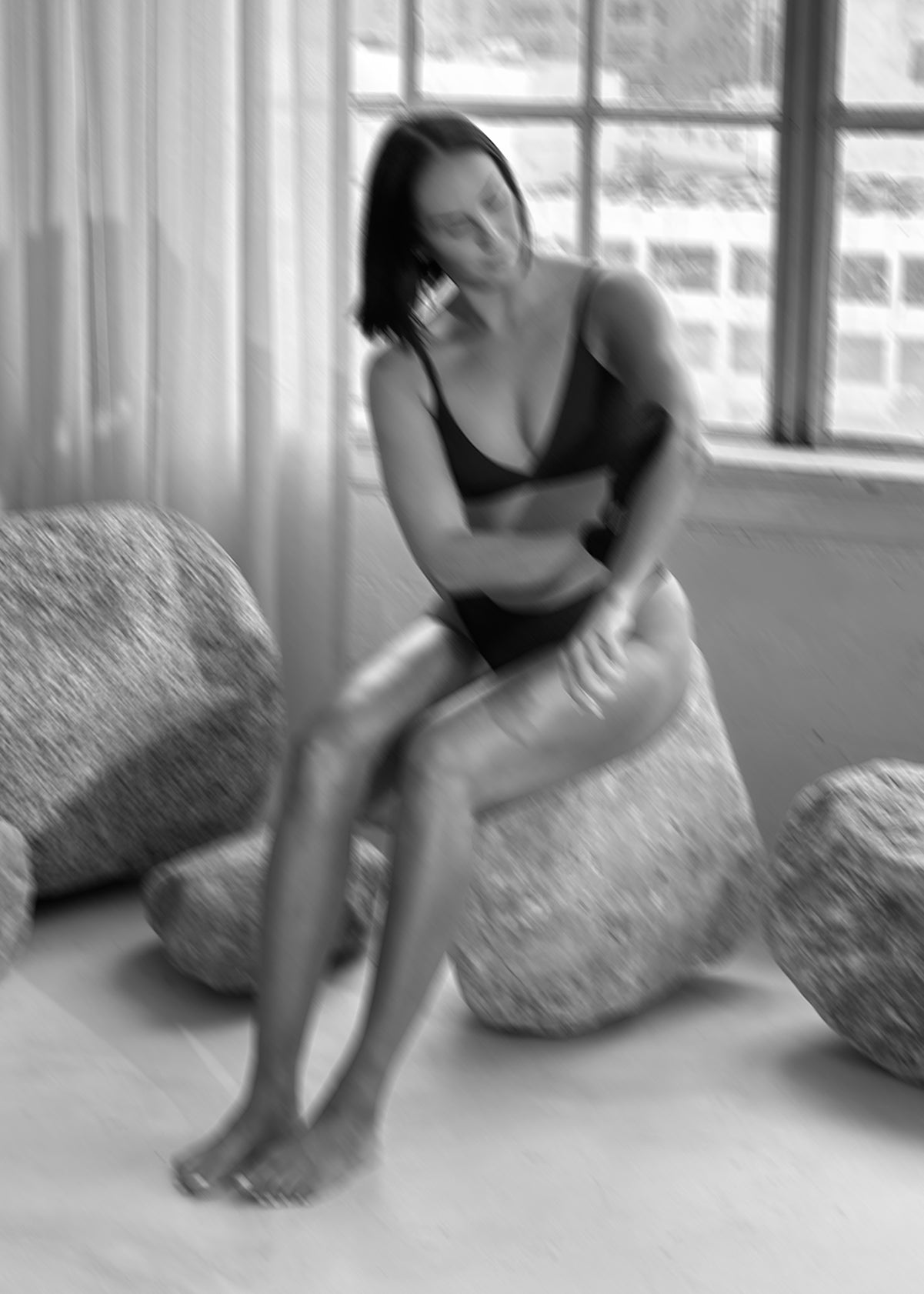 Woman in black bra and underwear seated on rounded stone-like cushions by a window, blurred black-and-white photo.