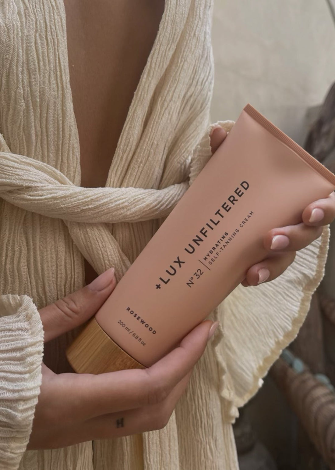 Front view of LUX Unfiltered number 32 Rosewood hydrating self-tanning cream tube held in both hands against a cream textured garment