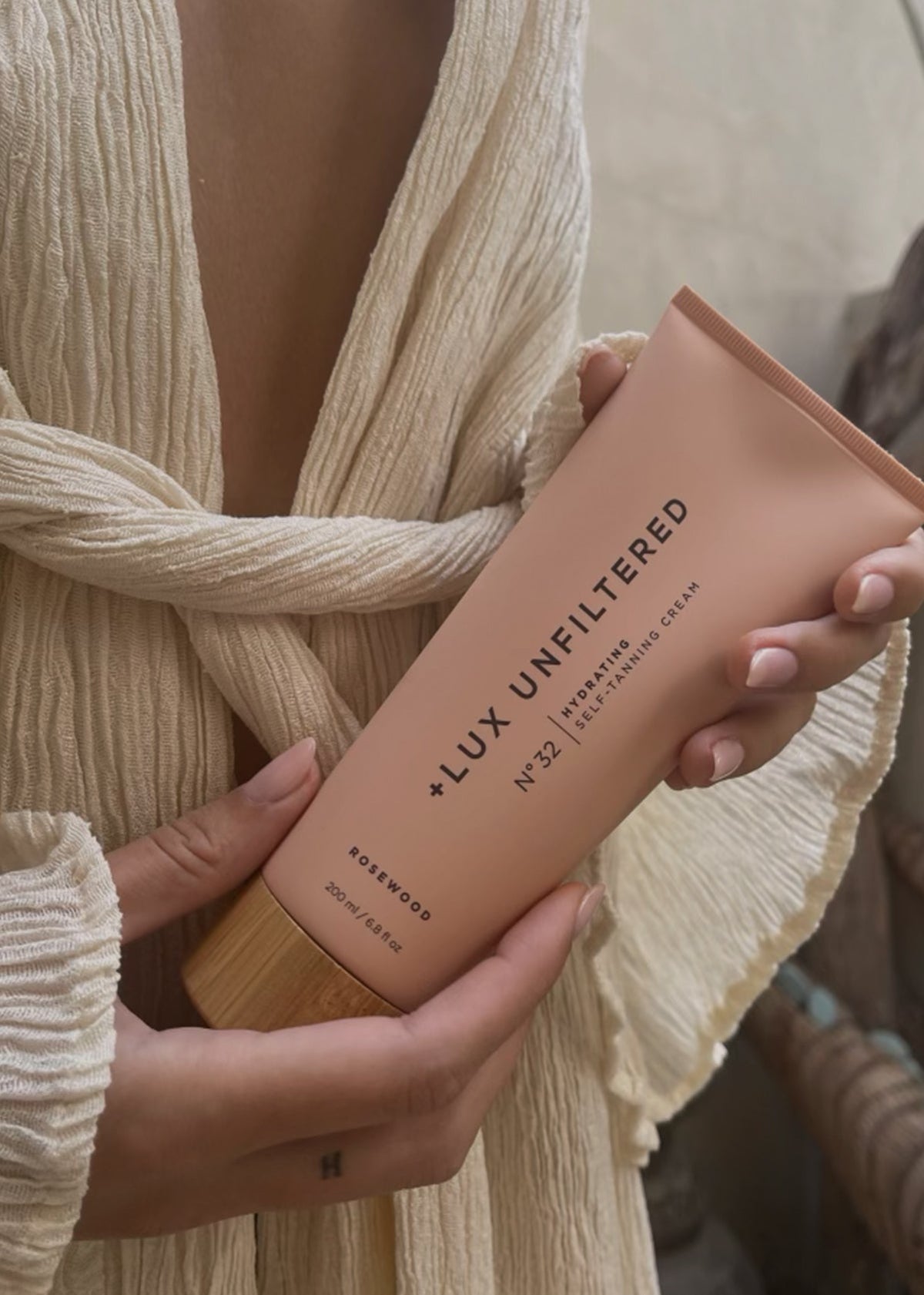 Front view of LUX Unfiltered number 32 Rosewood hydrating self-tanning cream tube held in both hands against a cream textured garment