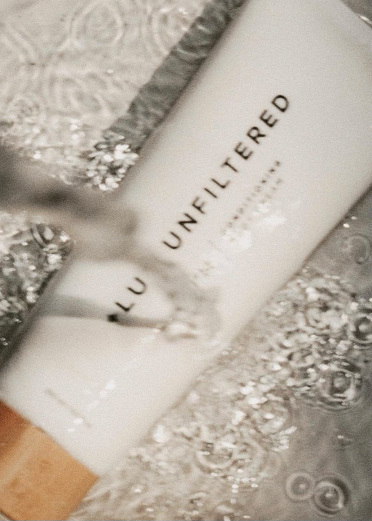 Close-up of a cosmetic tube labeled UNFILTERED with visible 'LU', resting on beaded fabric with water droplets.