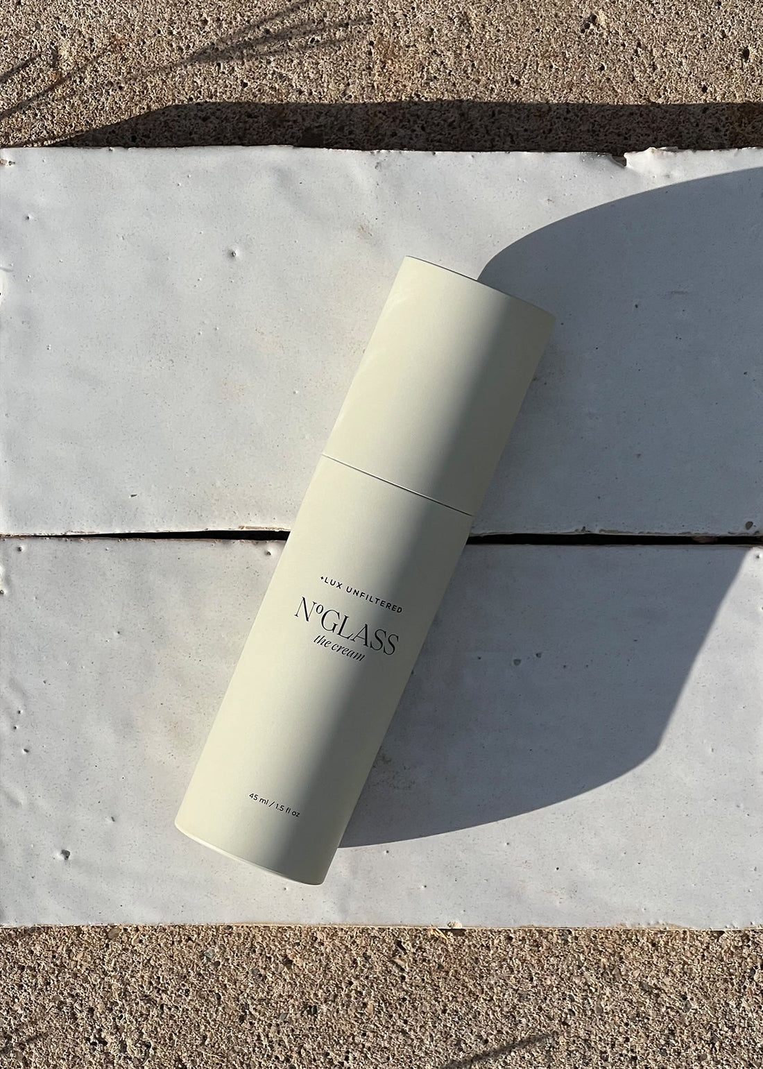 Pale cylindrical bottle labeled 'N°GLASS the cream, LUX UNFILTERED, 45 ml / 1.5 fl oz' lying on white tiles with shadow.