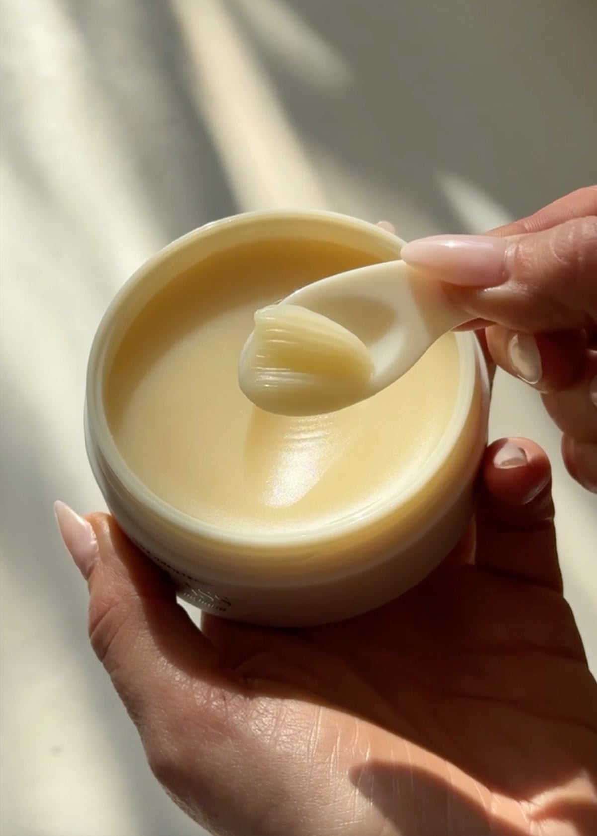 Pale-yellow cleansing balm in an open jar; small spatula lifts a scoop while hands hold the jar in soft sunlight.