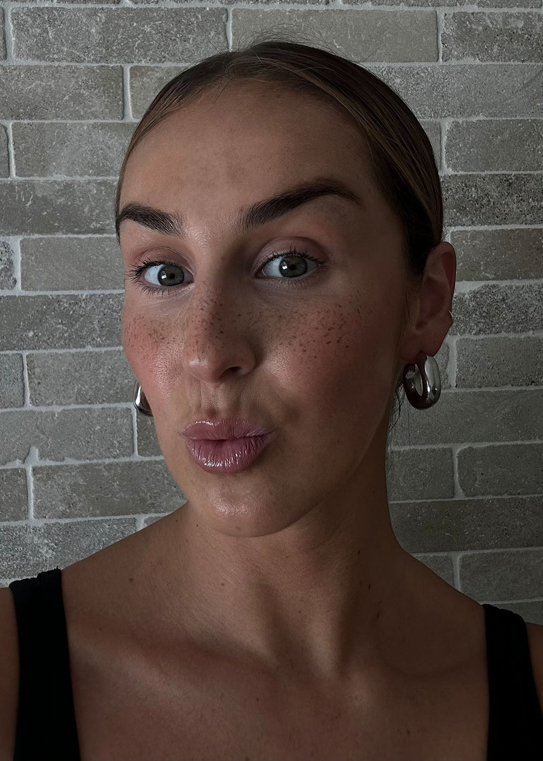Woman selfie showing mousse freckles across nose and cheeks, glossy lips, silver hoop earrings, brick-tile background.