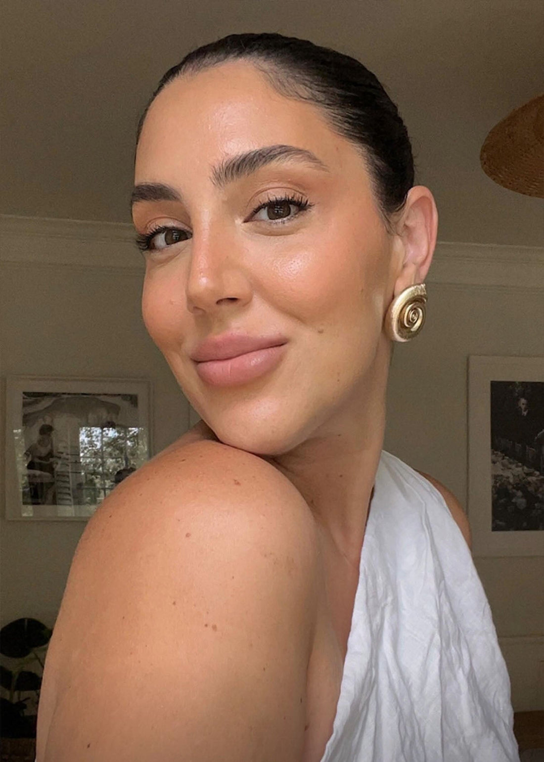 Head-and-shoulders portrait of a woman smiling, wearing a white one-shoulder top and a gold spiral earring.