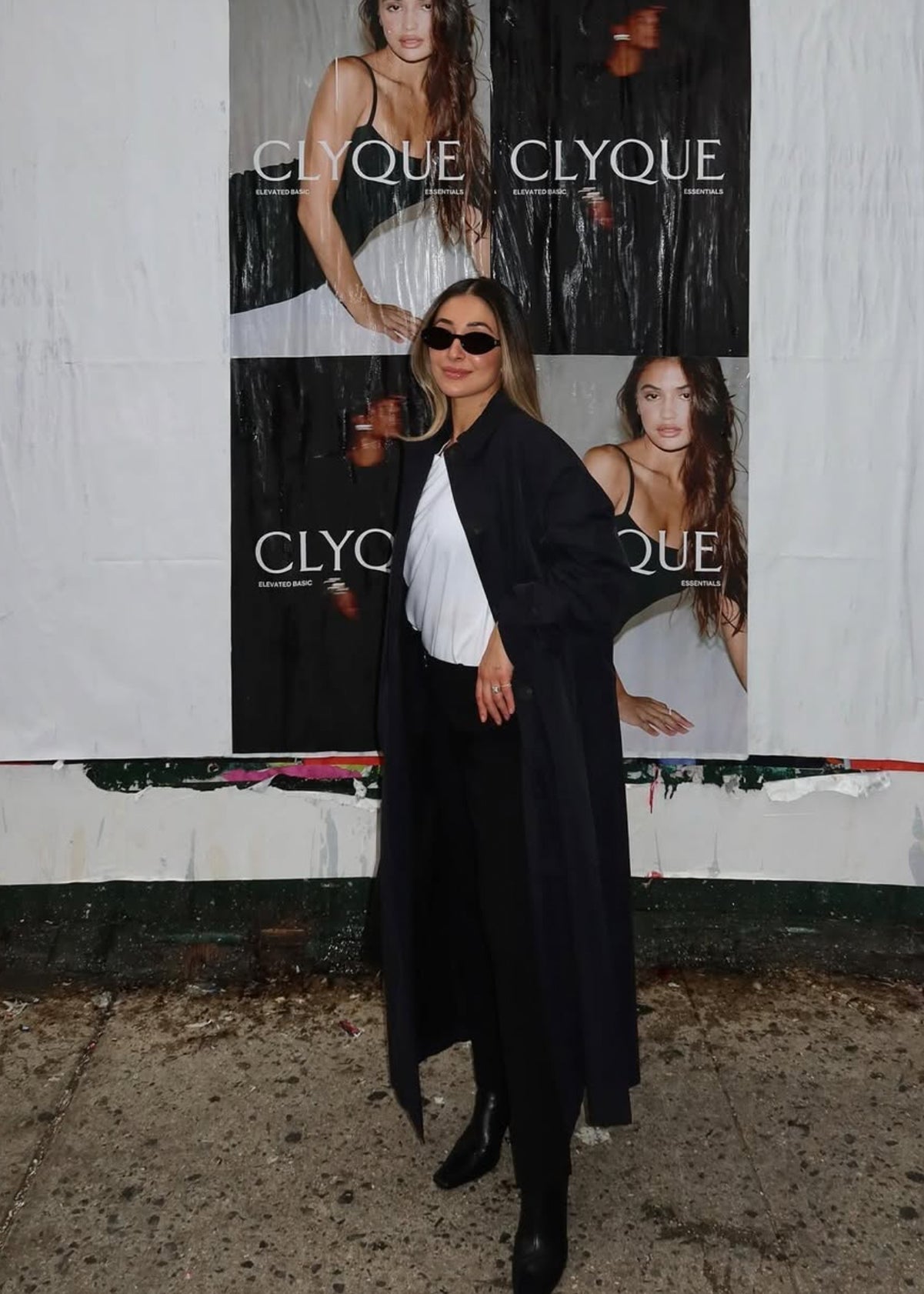 Woman wearing a long dark coat and sunglasses posing in front of posters reading CLYQUE, ELEVATED BASIC, ESSENTIALS