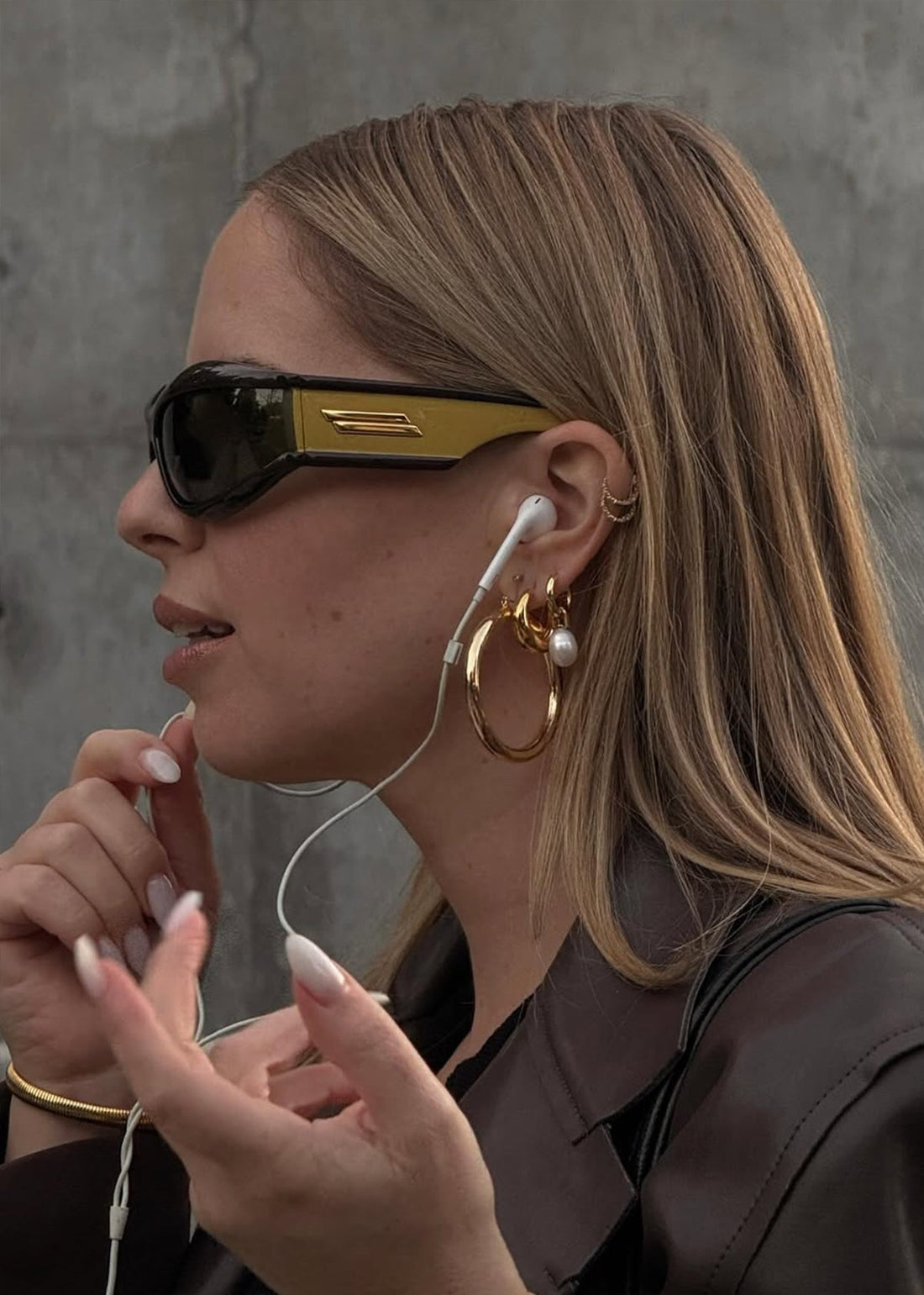 Side-profile of a woman wearing black sunglasses, a white earbud, and layered gold hoop earrings with a pearl, holding the cord.