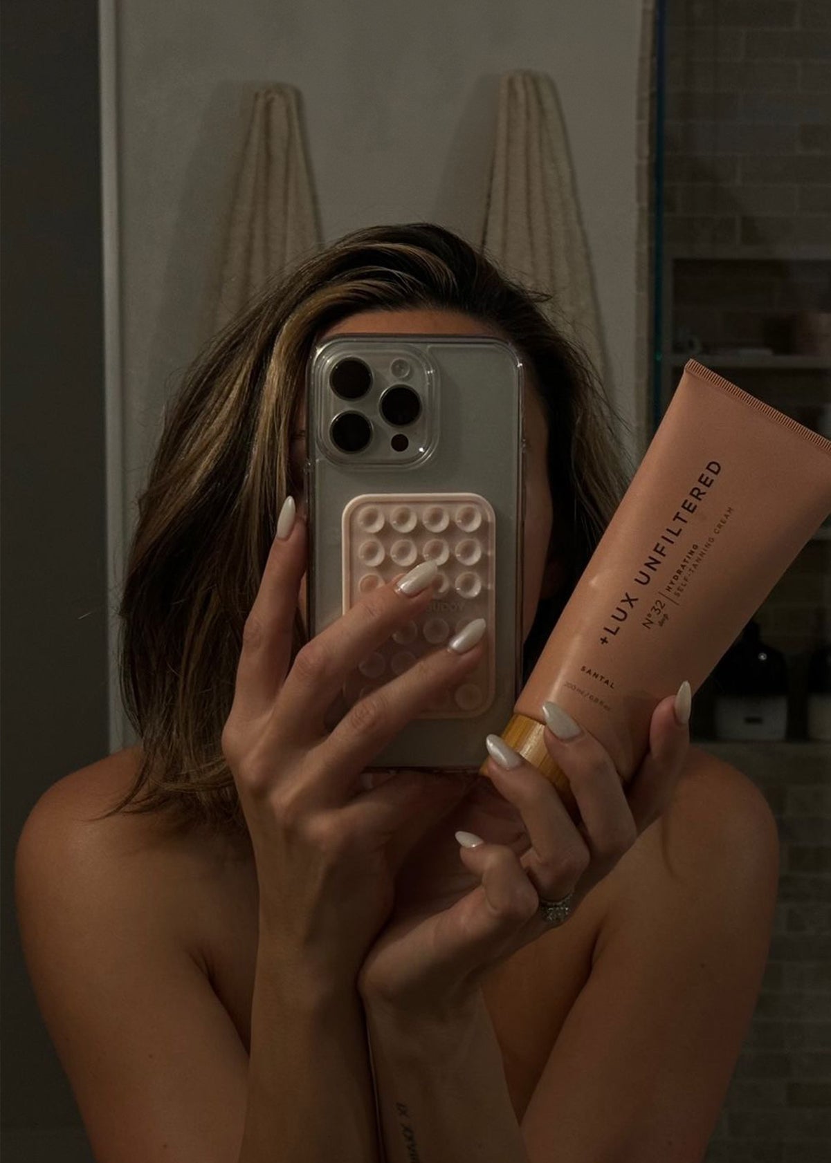 Mirror selfie: person holds phone covering face and a tube labeled '+LUX UNFILTERED N°32 HYDRATING SELF-TANNING CREAM SANTAL'.