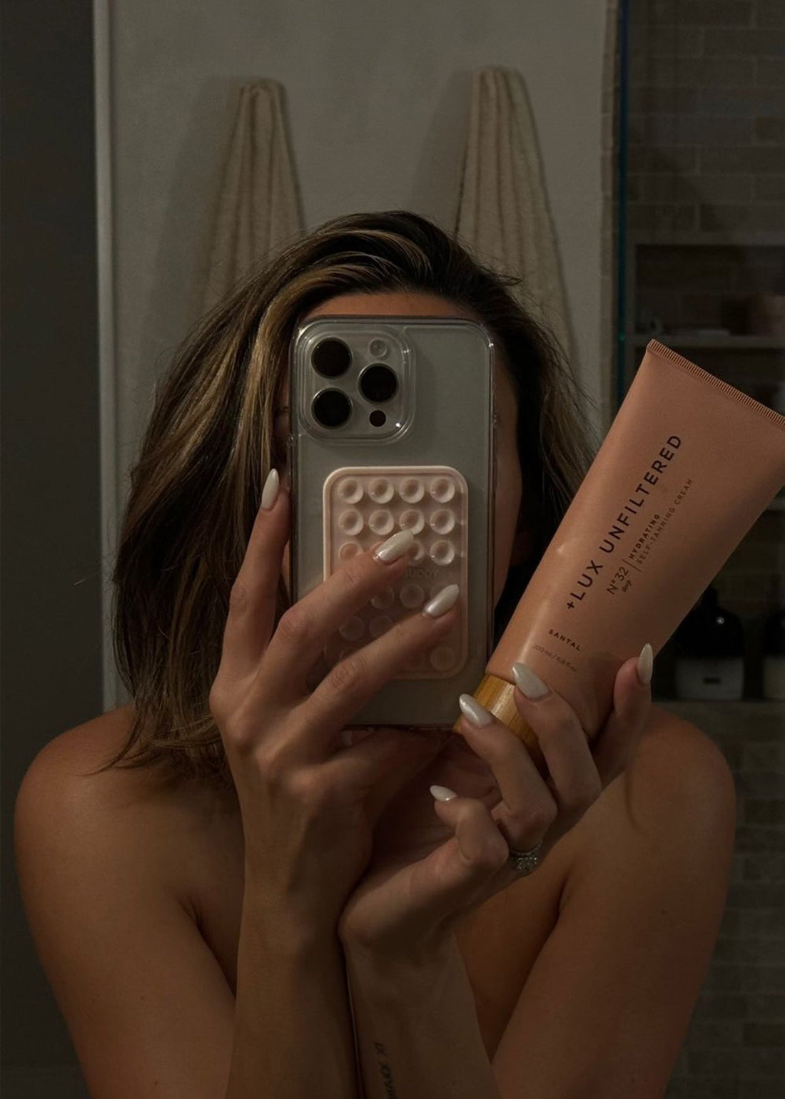 Mirror selfie: person holds phone covering face and a tube labeled '+LUX UNFILTERED N°32 HYDRATING SELF-TANNING CREAM SANTAL'.