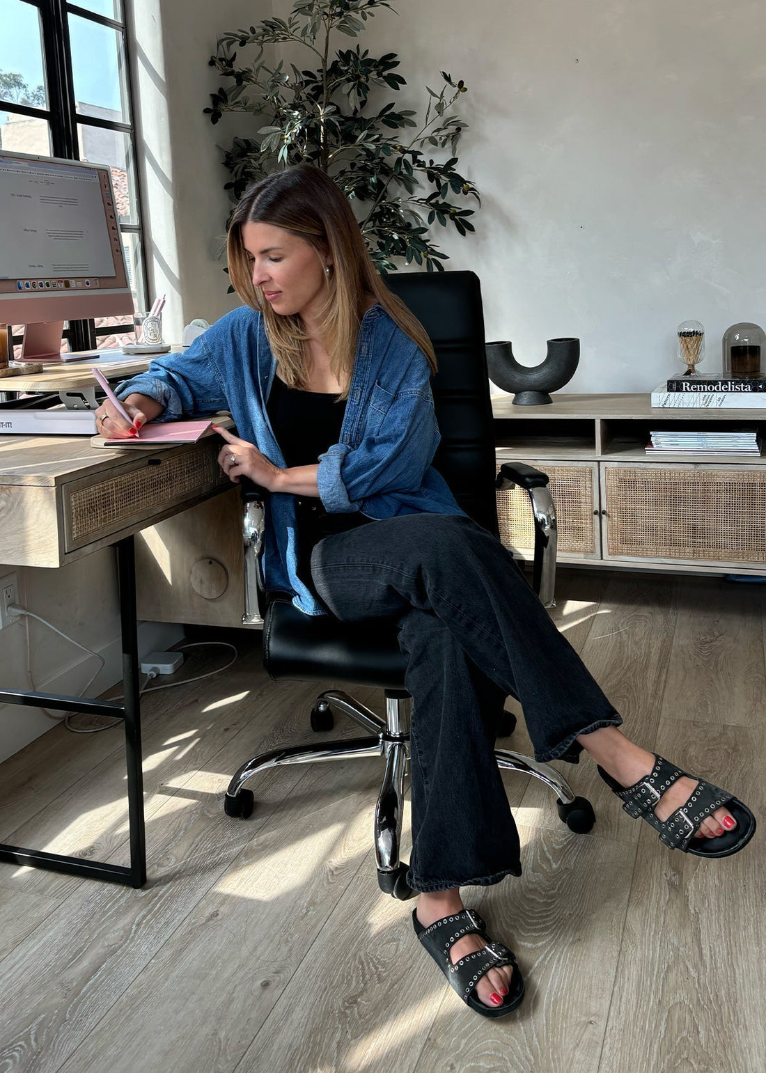 Woman at a home office desk writing in a notebook, denim shirt and black sandals; books on shelf include 'Remodelista'.