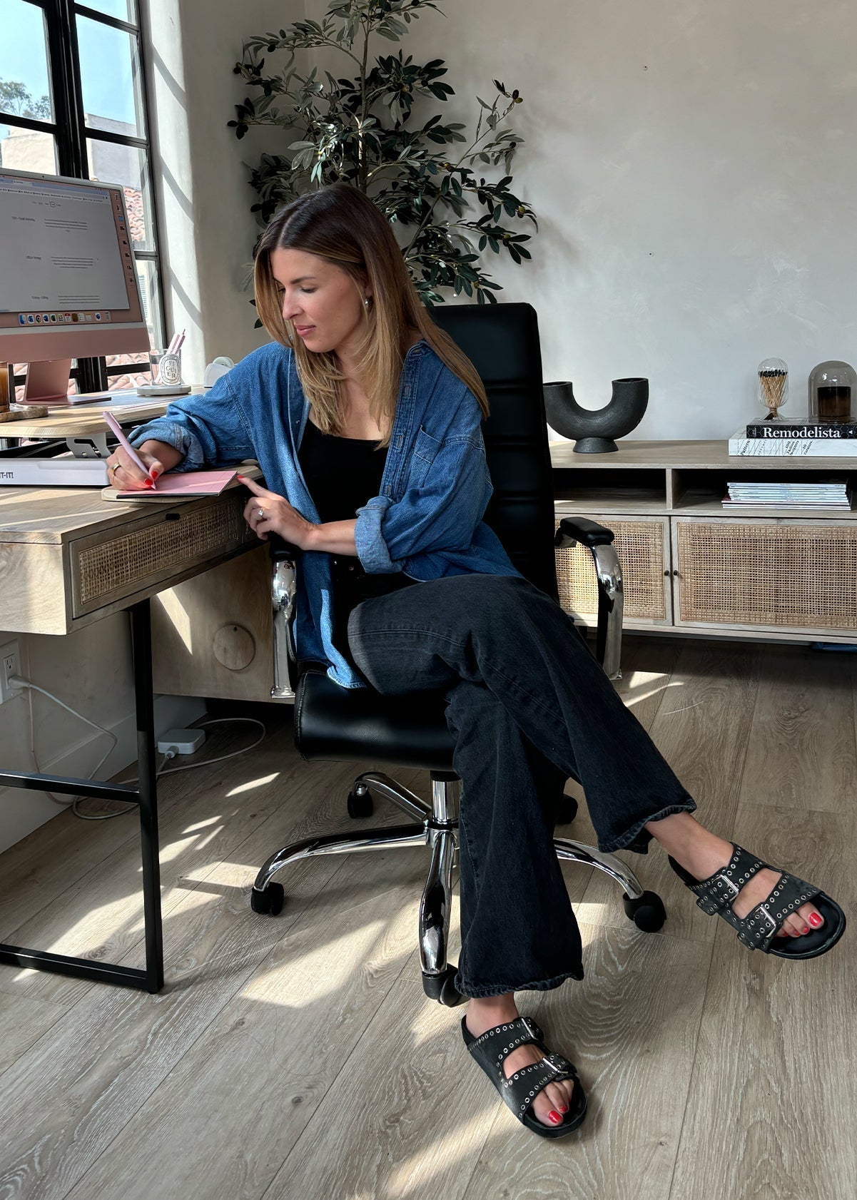 Woman at a home office desk writing in a notebook, denim shirt and black sandals; books on shelf include 'Remodelista'.