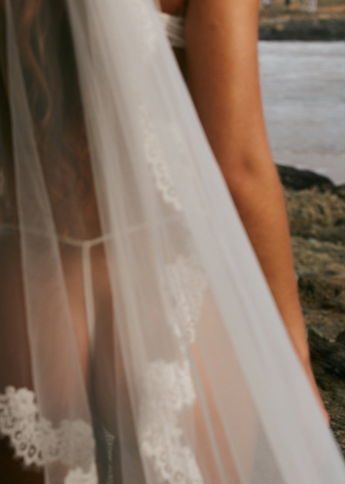 Sheer lace wedding veil draped over the bride's hips and arm with rocky shoreline in the background.