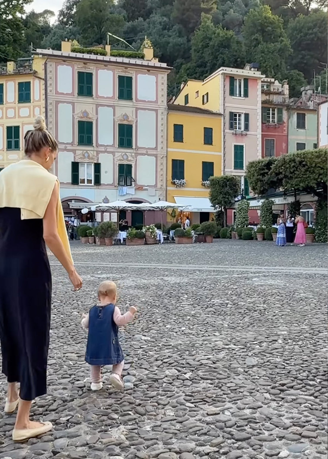 Woman walking with a toddler across a cobblestone square in Portofino, pastel buildings and cafés behind them.