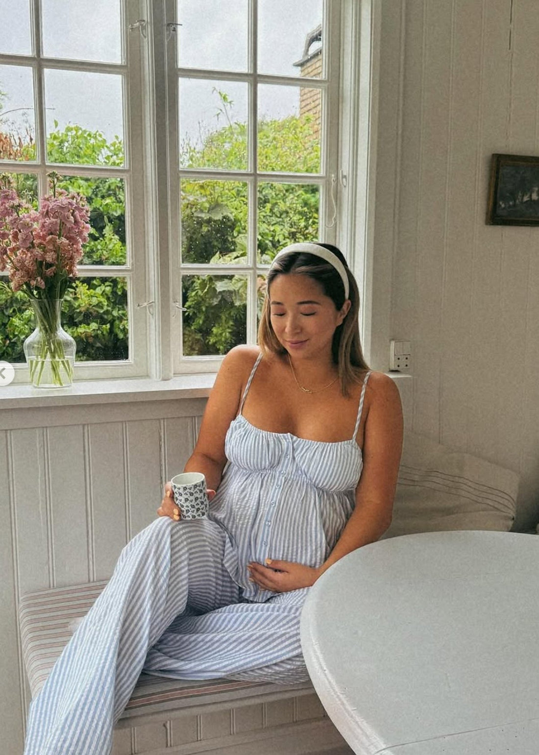 Pregnant woman in blue-striped dress sitting by a window, hand on belly, holding a patterned mug; pink flowers on sill