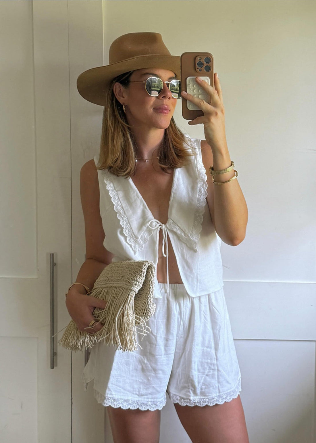 Woman mirror selfie wearing white linen eyelet top and shorts, tan hat, sunglasses, holding woven fringe clutch