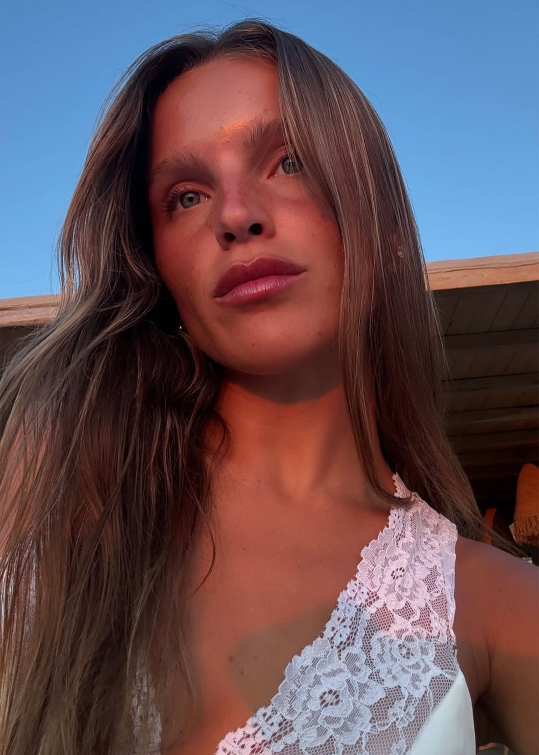 Young woman looking up, warm sunset light on her face, long hair and white lace top against blue sky