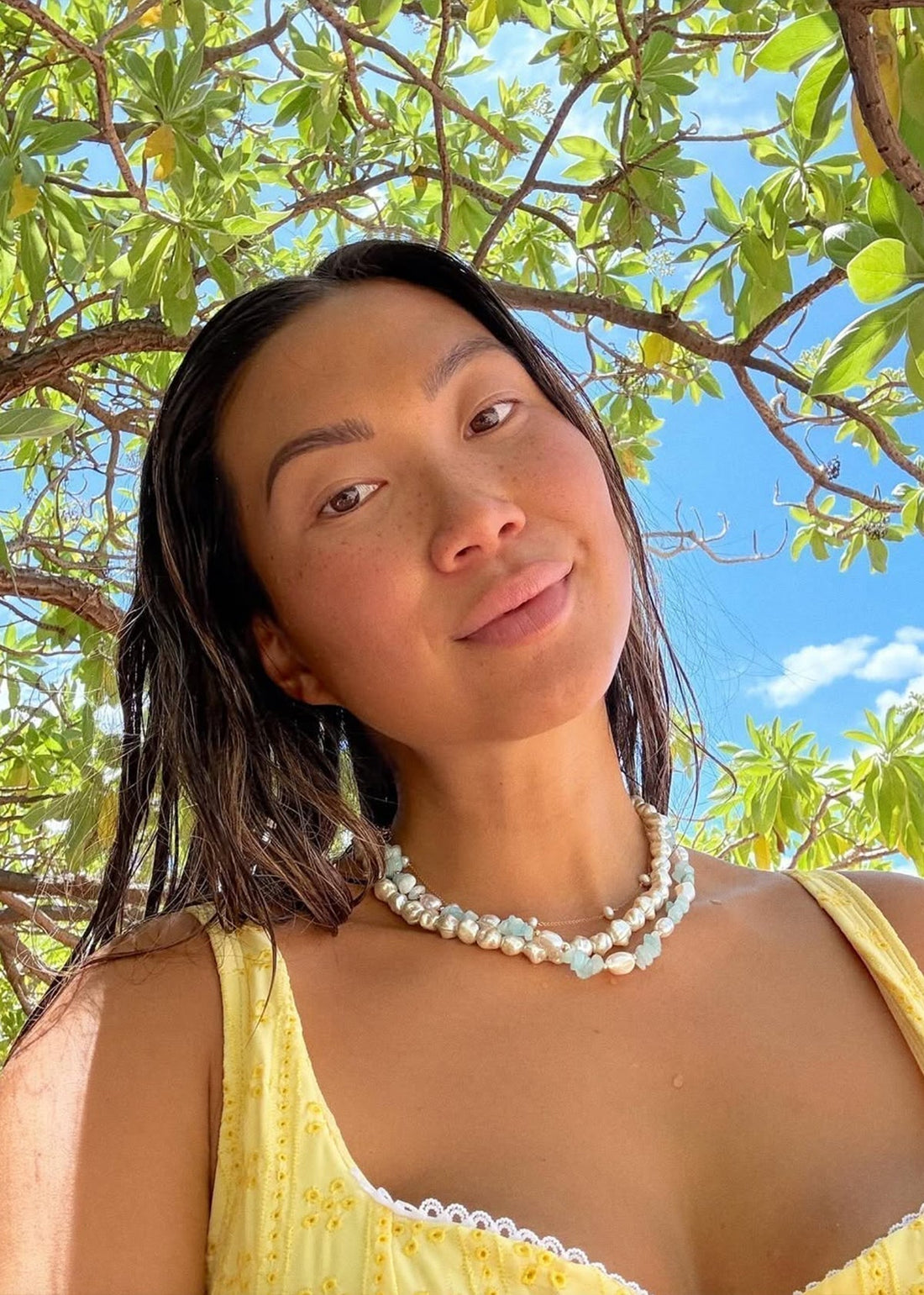 Selfie of a woman under a leafy tree and blue sky wearing a yellow sundress and layered pearl-and-aquamarine necklaces