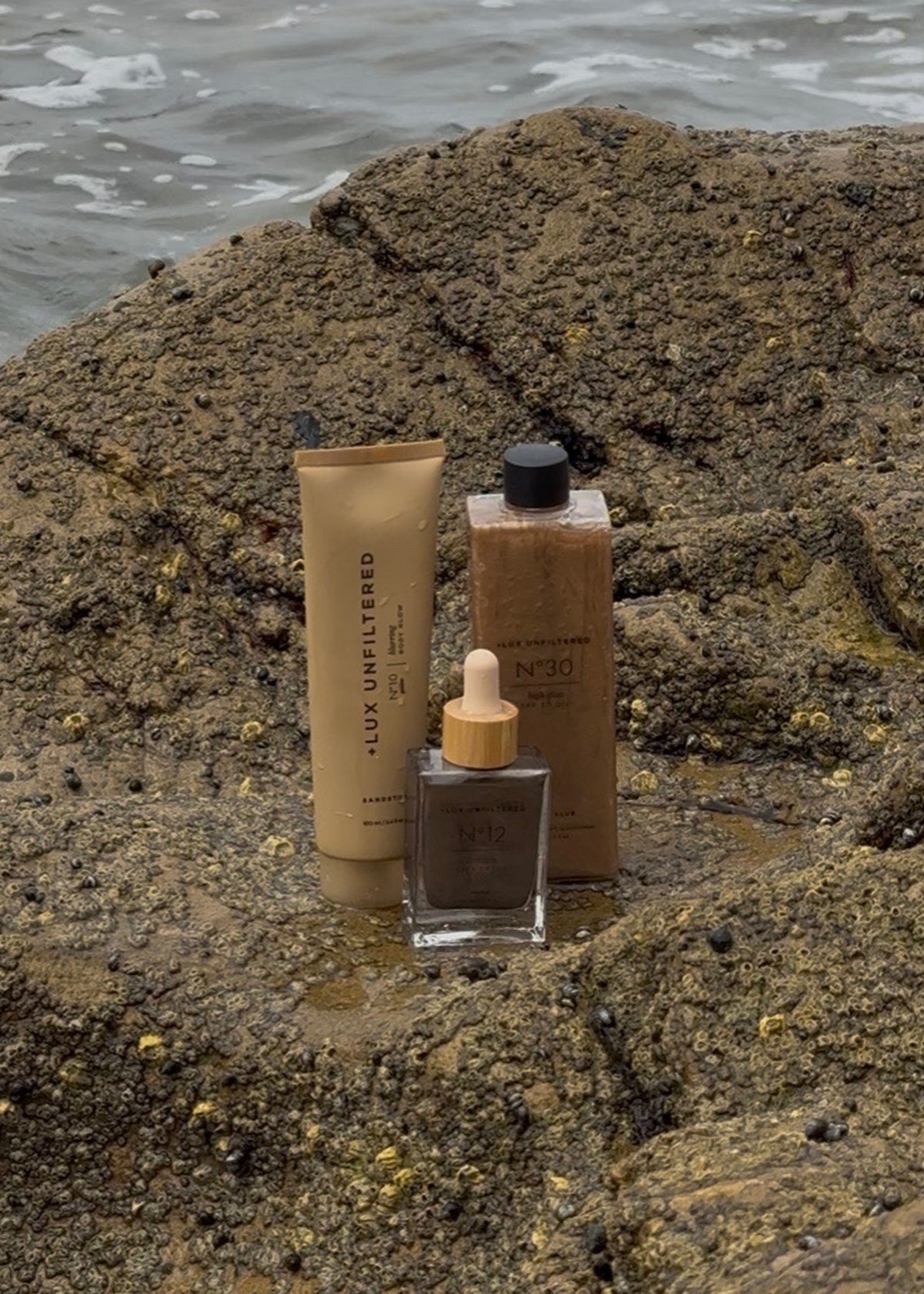 Three LUX Unfiltered skincare products, including a tube, capped bottle, and dropper bottle, displayed on barnacled rocks by the sea
