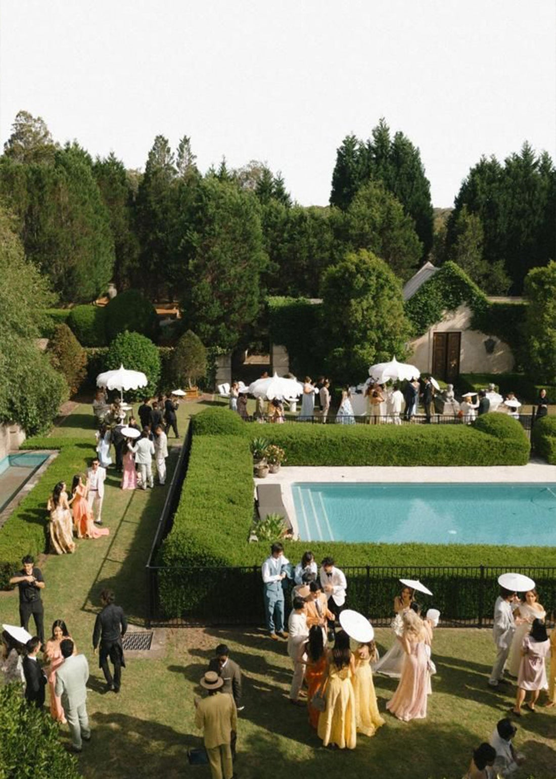 Wedding guests mingling by a rectangular pool and hedged garden, some holding white parasols, wearing pastel formal attire.