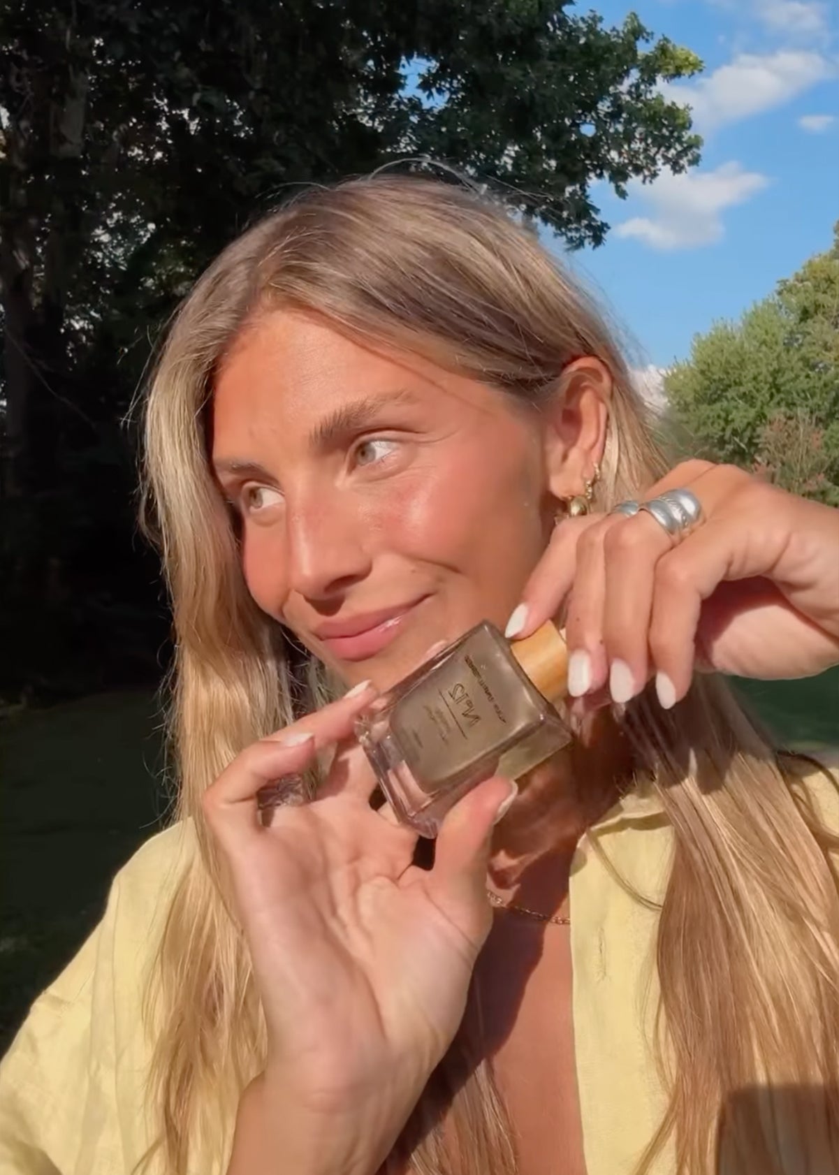 Woman outdoors smiling, holding a perfume bottle close to her face