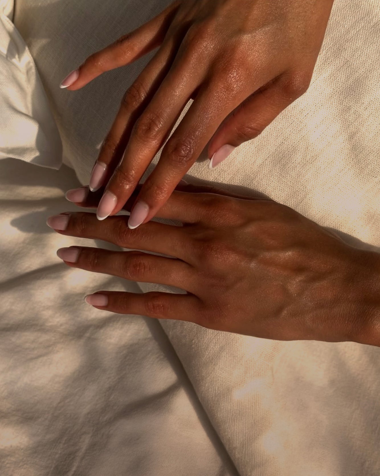 Hands with pale pink manicured nails resting on cream-colored bedding in warm light