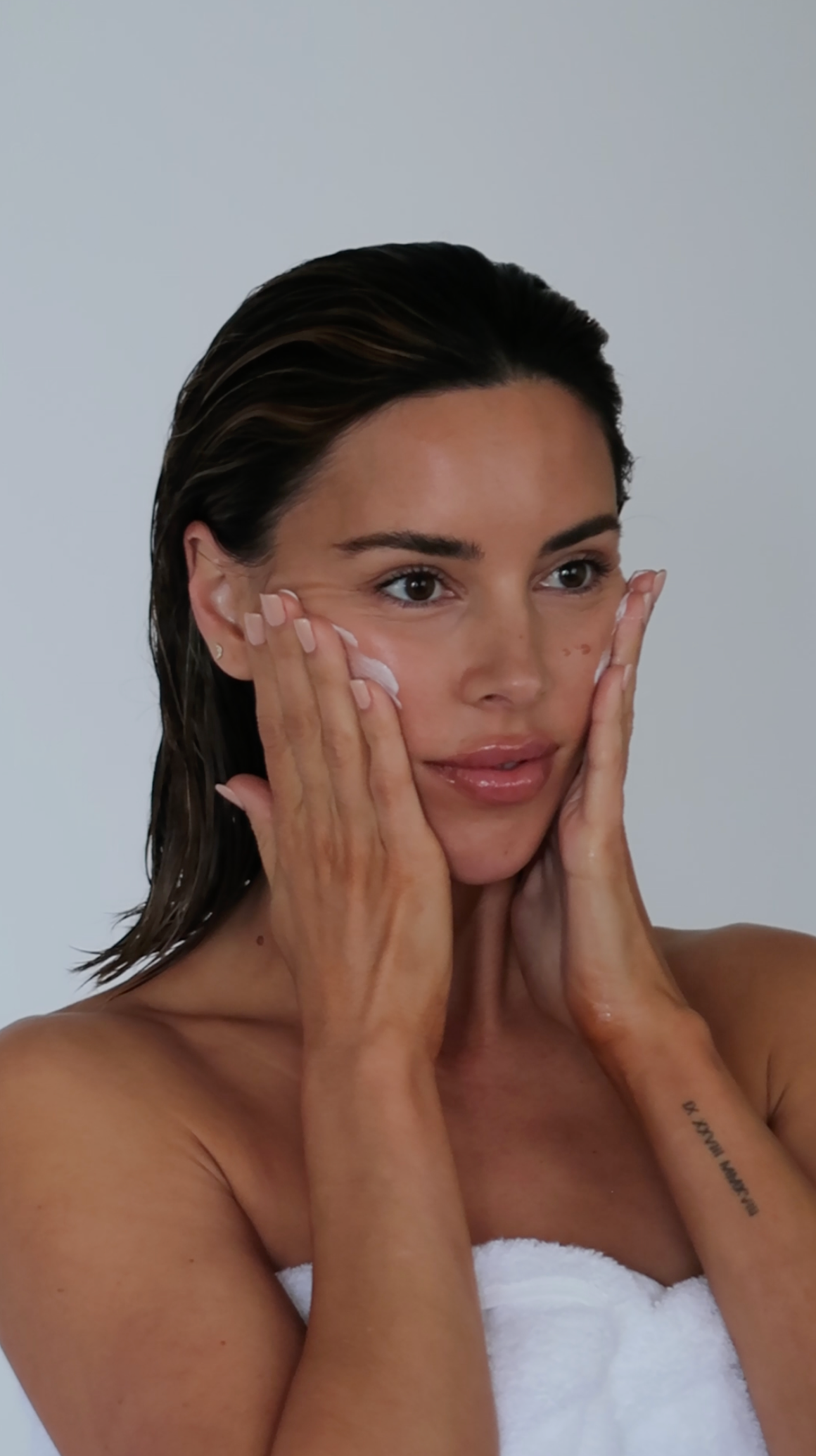Woman applying skincare cream to her cheeks while wrapped in a white towel against a plain background
