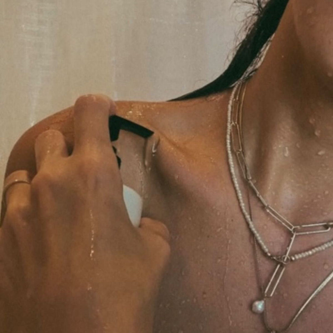 Close-up of a person spraying self-tanning mist onto a wet shoulder in the shower, layered silver necklaces visible