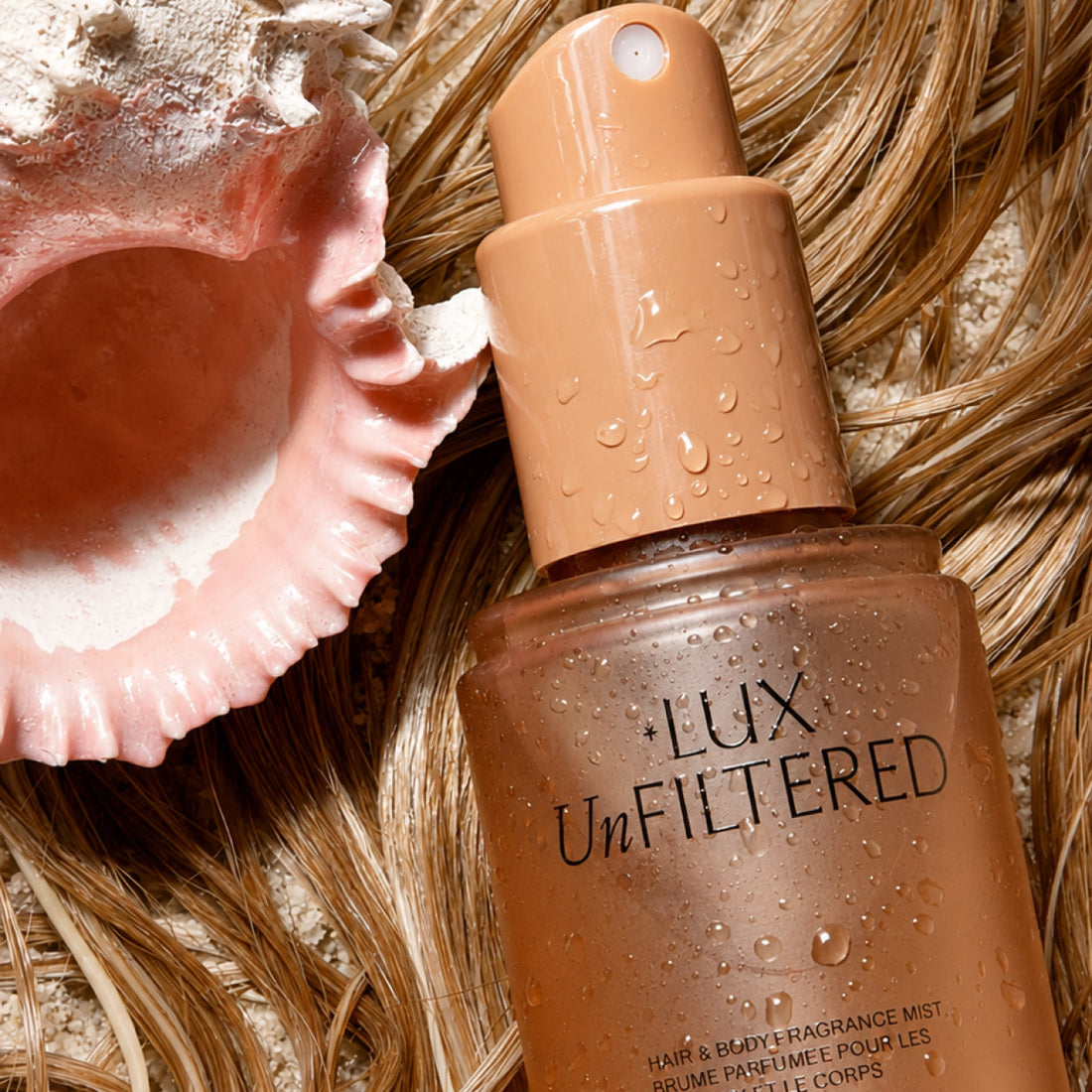 Close-up of LUX Unfiltered hair and body fragrance mist bottle with water droplets on textured hair beside a seashell