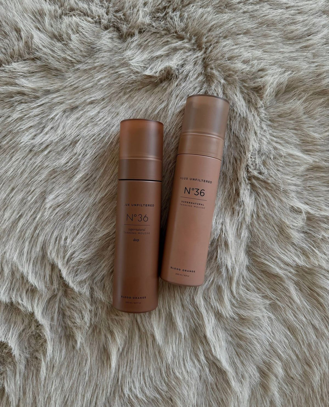 Top view of two LUX UNFILTERED N°36 Supernatural Tanning Mousse bottles, including the deep shade, on gray faux fur