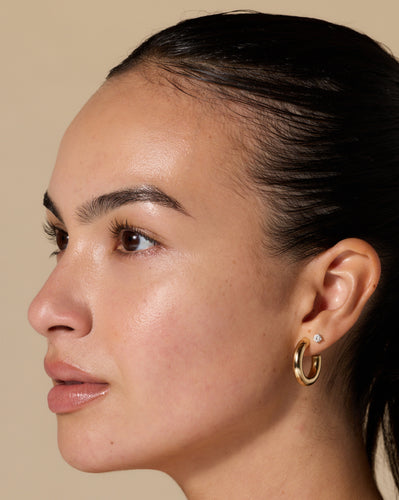 Close-up profile of woman wearing gold hoop and small diamond stud earring, dewy skin and slicked-back hair.