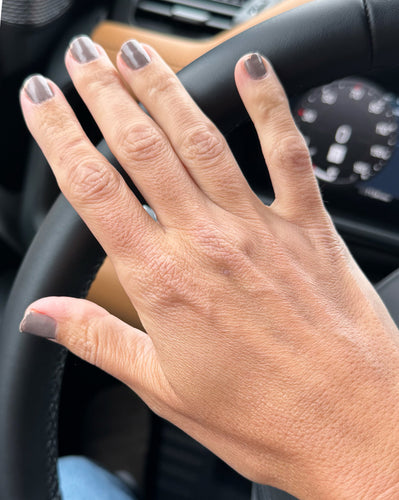 Left hand with taupe-gray nail polish resting on a steering wheel; instrument cluster blurred in background.