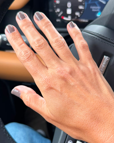 Left hand with taupe-gray nail polish resting on a car steering wheel; tan leather interior and dashboard visible.