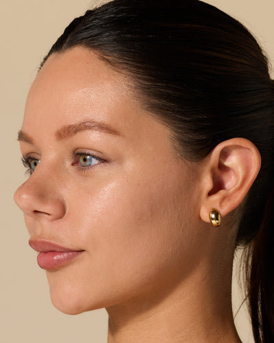 Close-up profile of a woman with slicked-back hair wearing a small polished gold hoop earring on her right ear.