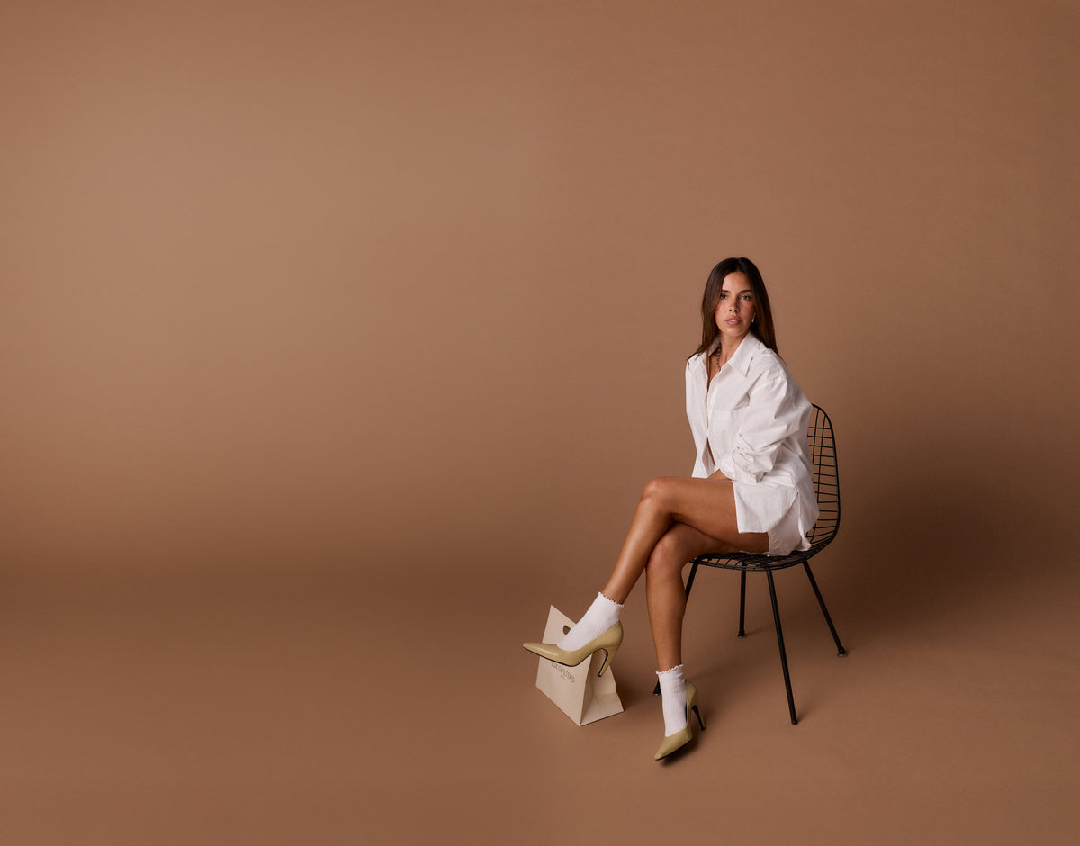 Woman in white shirt, socks and heels seated on wire chair against brown backdrop; crossed legs and bag at her foot.