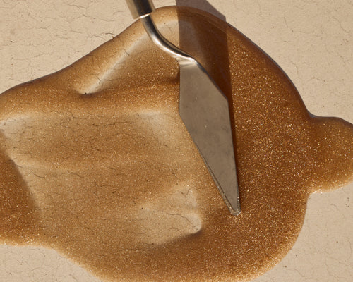 Close-up of High Gloss Body Oil showing golden shimmery texture with a metal spatula on a beige surface
