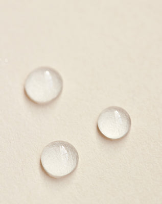 Close-up of Après Sun Tan Extending Serum droplets showcasing a lightweight hydrating gel texture on a neutral beige background