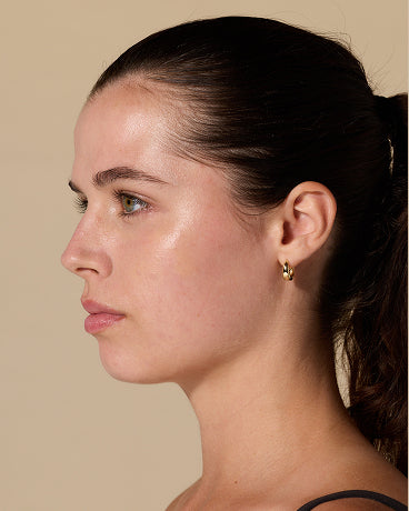 Young woman in left-facing profile with dark hair in a ponytail and a small gold hoop earring against a beige background.