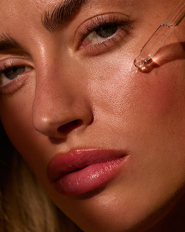 Close-up of a woman's face with dewy skin as a glass serum dropper releases a droplet onto her cheek