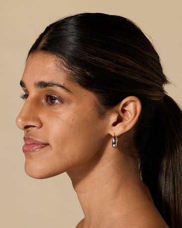 Profile of a woman with dark hair in a low ponytail wearing a small silver hoop earring on a beige background.