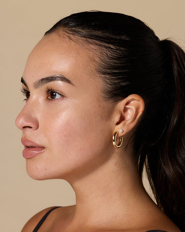 Profile of a woman with a ponytail wearing two gold earrings (hoop and huggie) on a beige background.