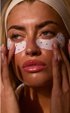 Woman's face close-up wearing white under-eye patches with small black crosses, glossy lips, manicured nails touching them