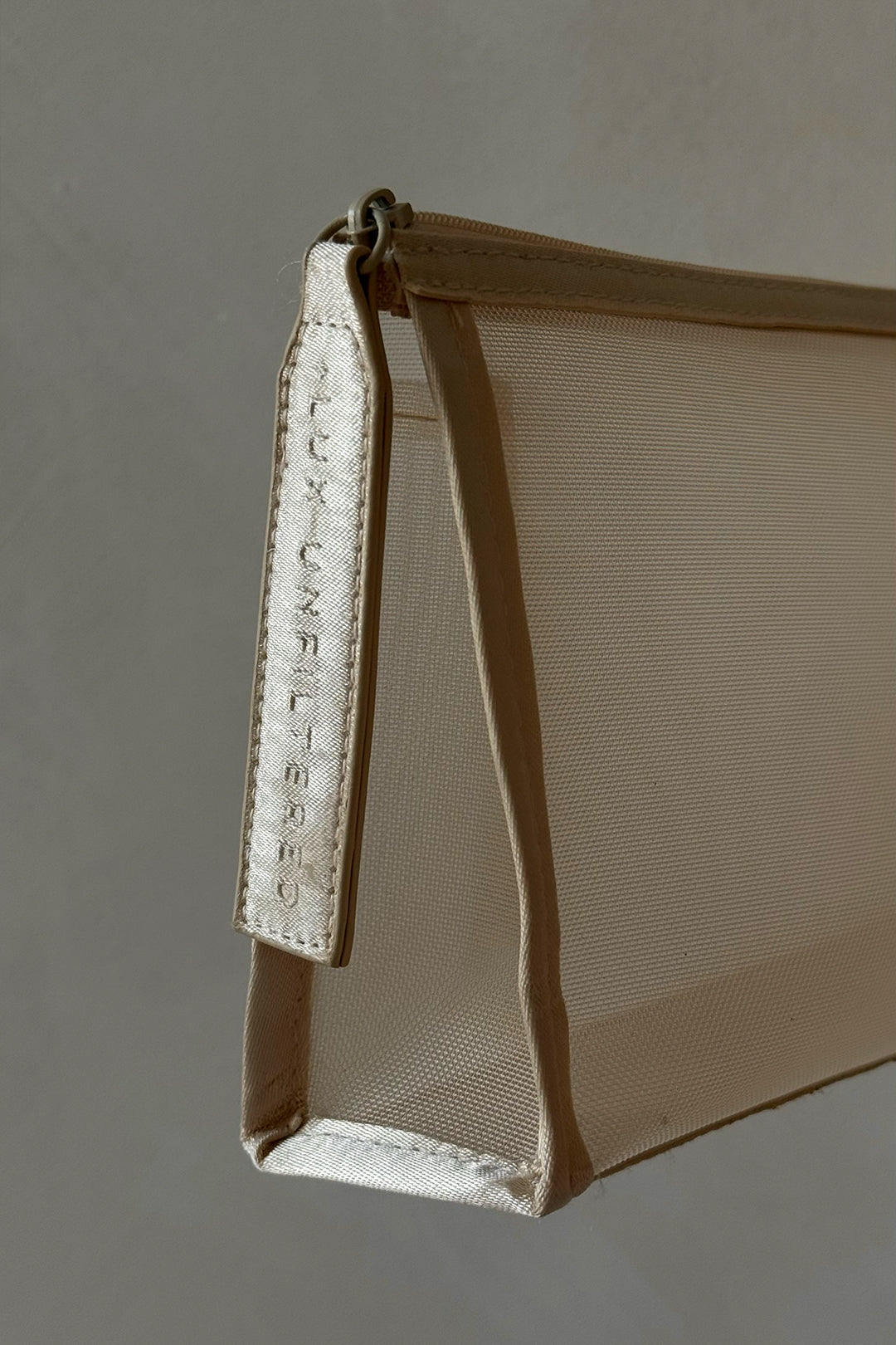 Beige mesh zip pouch with a strap debossed 'LUX UNFILTERED', photographed hanging.