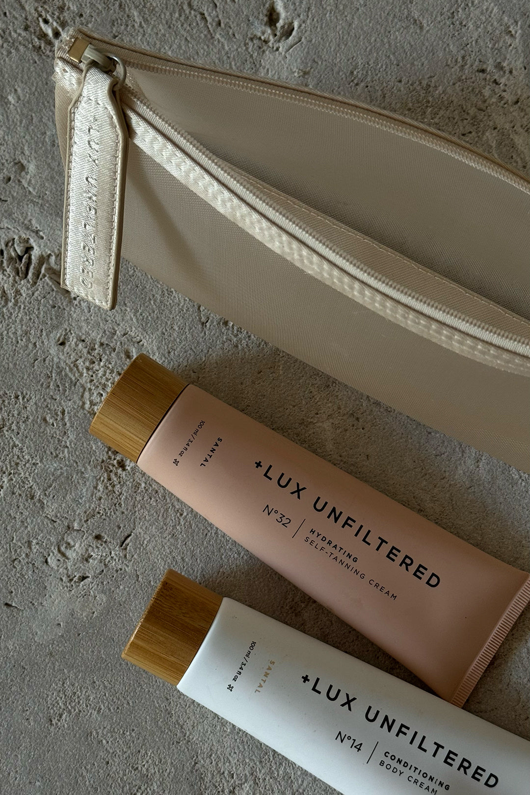 + LUX UNFILTERED tubes N°32 (hydrating self-tanning) and N°14 (conditioning body) with bamboo caps beside beige pouch on stone