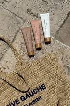 Three + LUX UNFILTERED tubes on stone next to a woven RIVE GAUCHE SAINT LAURENT straw bag.