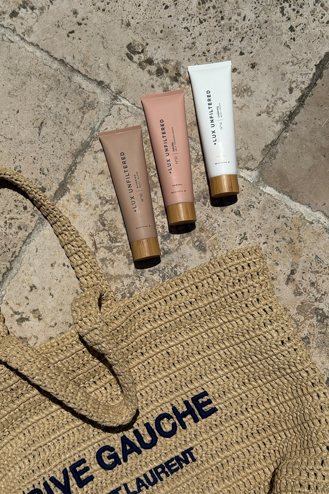 Three + LUX UNFILTERED tubes on stone next to a woven RIVE GAUCHE SAINT LAURENT straw bag.