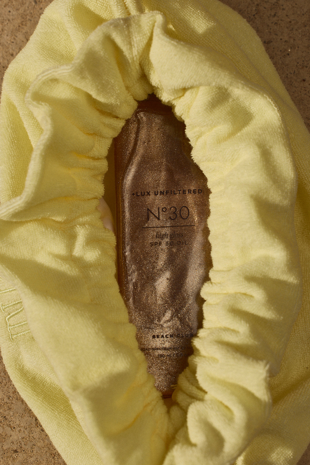 Yellow terry towel wrapped around bronze bottle labeled LUX UNFILTERED Nº30 high gloss SPF 30 OIL BEACH CLUB