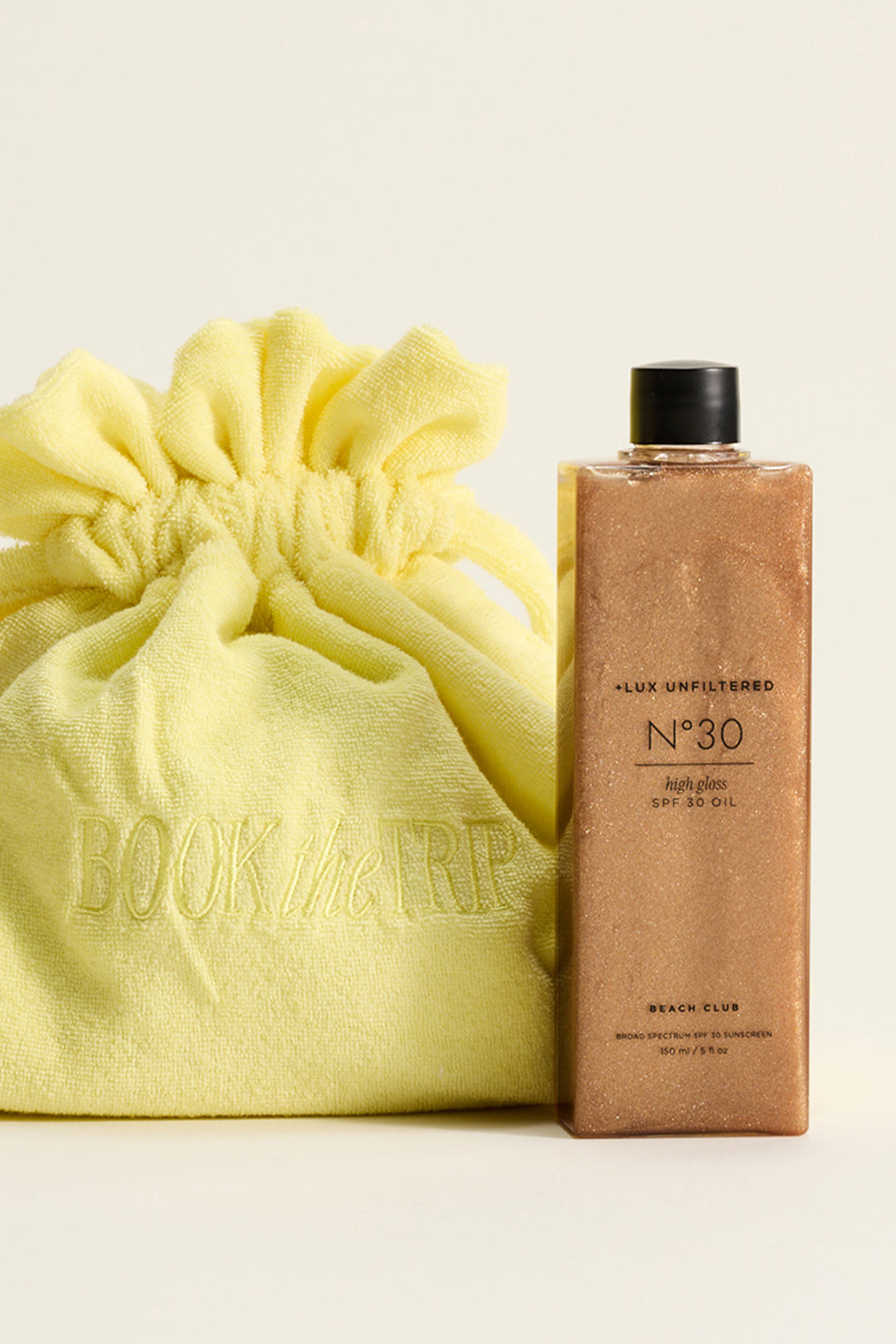 Bottle labeled +LUX UNFILTERED N°30 high gloss SPF 30 OIL BEACH CLUB beside yellow drawstring bag embroidered BOOK the RP